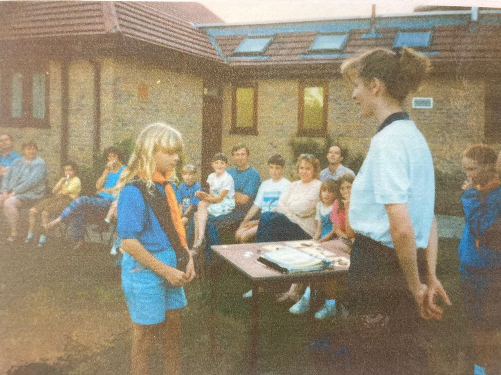 Making my Promise as a Guide, outside in the summer. I'm wearing denim shorts, the blue Jeff Banks polo shirt with a saffron neckerchief and bare sash and there are various parents and siblings watching the ceremony. The photo is a scan of a 35mm film photo, so it's not great quality.