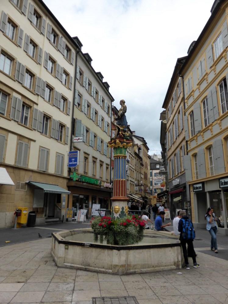 The yellow streets of central Neuchatel. In the middle is a tall colourful fountain.