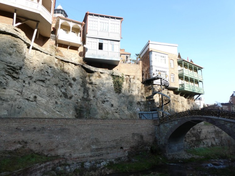 Houses and balconies hanging over a relatively low cliff towering over the brick sides of the shallow gorge below. Most of them are elaborate balconies and there's a metal spiral staircase leading up to the top of the cliff.