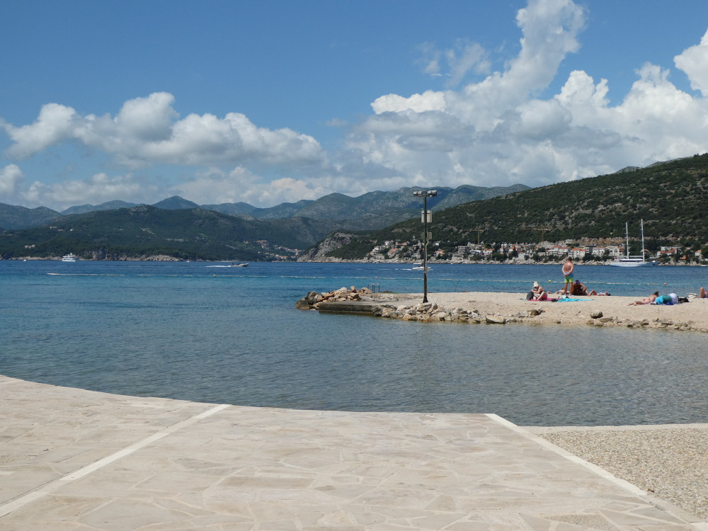 Copacabana Beach, a rounded bay of bright blue water, stone slabs along the edge and sand on the peninsula opposite. On the other side of the bay is the entrance to Dubrovnik's port and the steep mountains of the coastline.