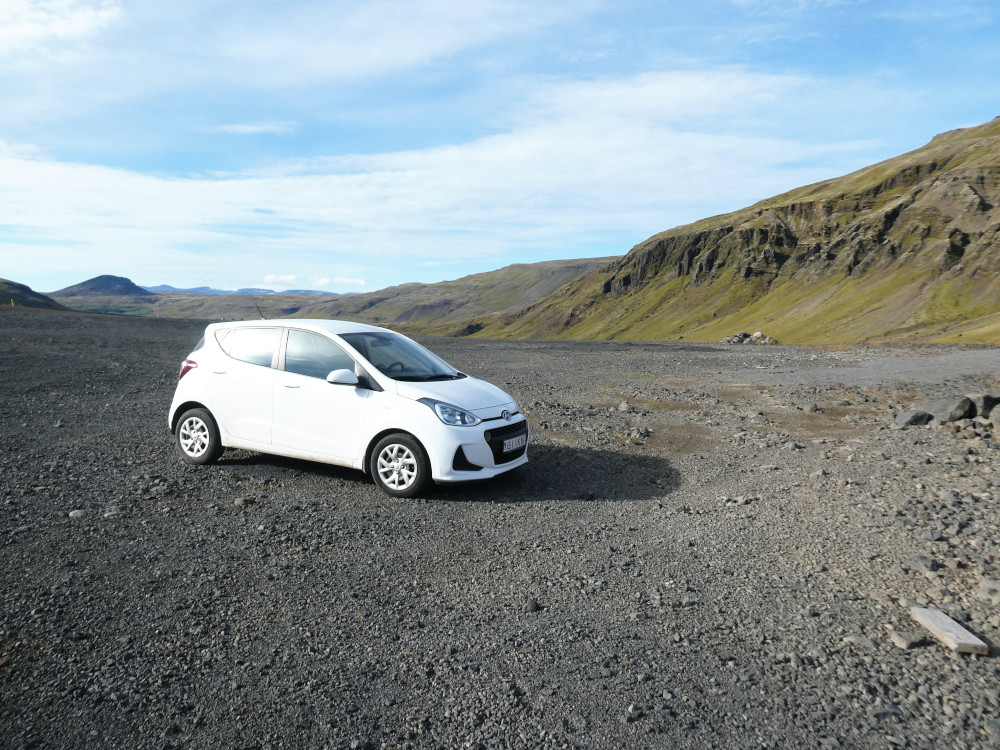 A white Hyundai i10 parked on a big patch of gravel. To the right is a craggy moss-streaked mountain and in the background are some slightly bigger mountains. The sky is more blue than cloud.