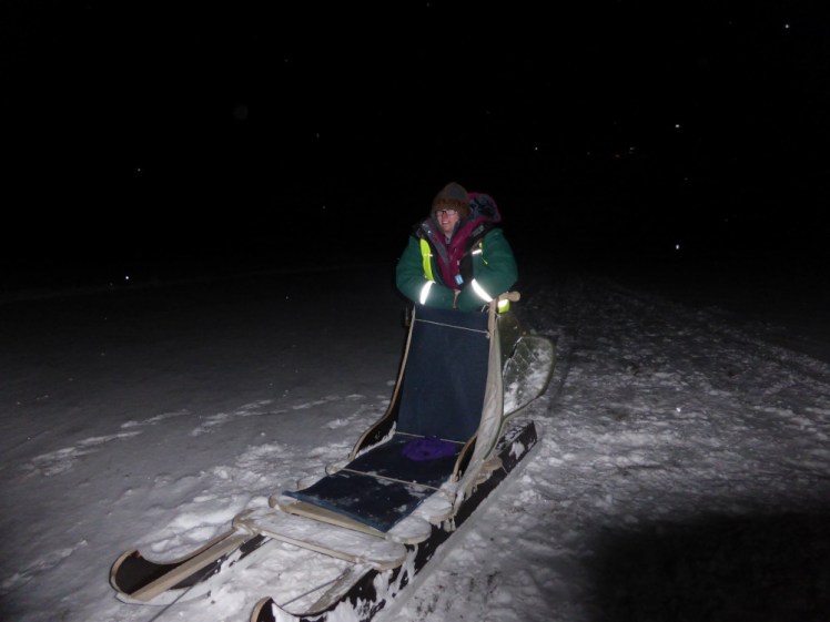 Me, bundled up in lots of layers, hanging onto the back of a dogsled. The dogs are not harnessed to it - we couldn't take photos in action so this is posing with the sled afterwards. It's the polar night in Svalbard and I'm in a spotlight of a camera flash.