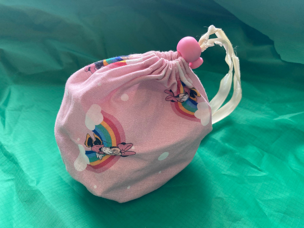 A small drawstring bag made of pink fabric with Minnie Mouse and rainbows. It's held together by a cream ribbon and a matching pink toggle.