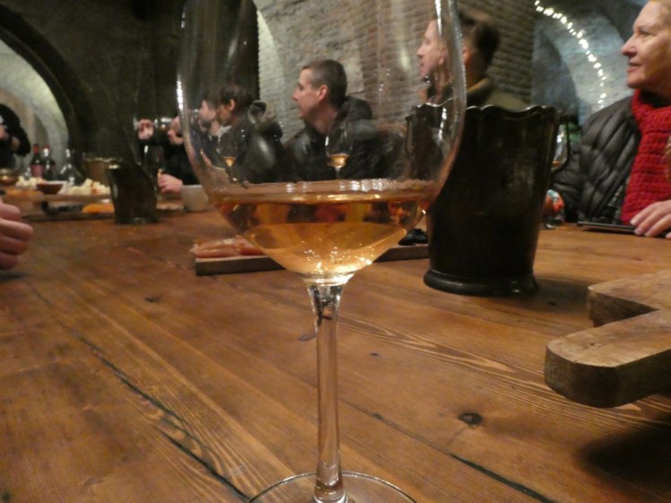 A glass of amber white wine on a large wooden table. Along the sides of the table are lots of content creators.