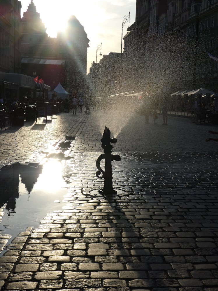 An old-fashioned water pump in the cobbled square spraying out water from the top. Pump and water are silhouetted by the low sun behind them.