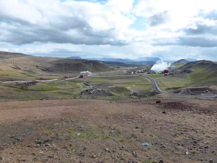 A view down into a fairly wide valley which is criss-crossed with heavy-duty pipes, red geodesic domes housing boreholes and red power station buildings issuing clouds of steam.