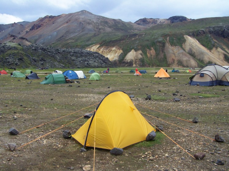 A small yellow tent set up in a field that's more gravel than grass, with a mossy lava field and volcano range in the background.