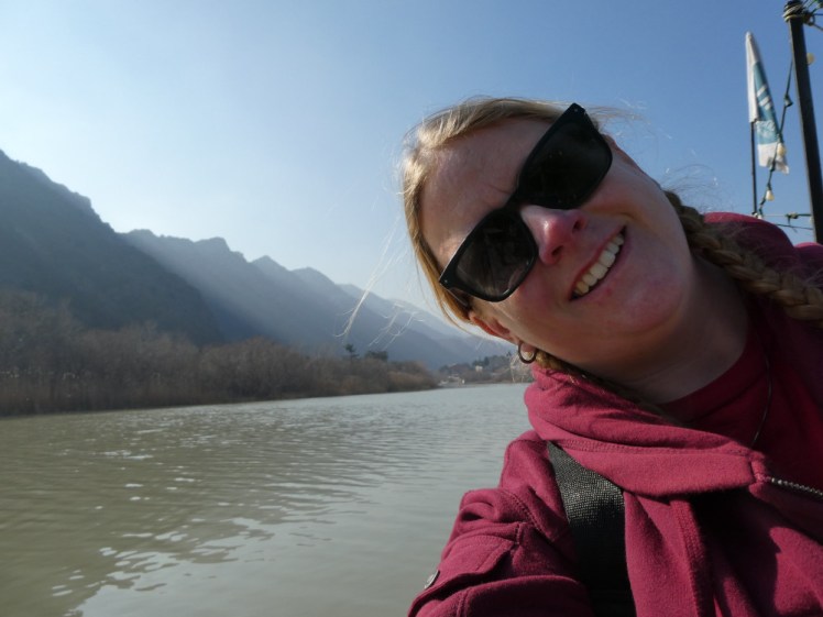 A selfie down by the river in Mtskheta. I'm wearing a burgundy hoodie with a red t-shirt and the sun is right behind me. There are mountains on the other side of the river, which is kind of brown but otherwise very beautiful.