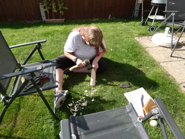 Me, sitting on the grass in the back garden, carving a spoon out of a chunk of wood with my axe