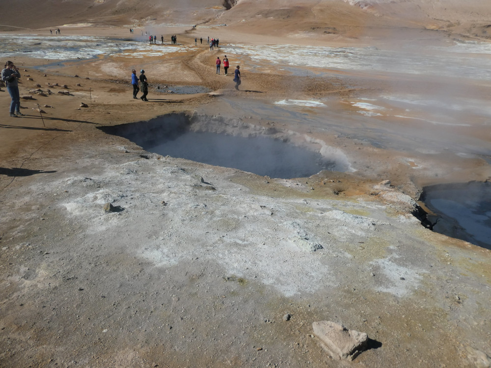 Dark holes in the orange-white landscape filled with bubbling dark blue mud and steaming. Tourists walk between them on scraped-out paths.