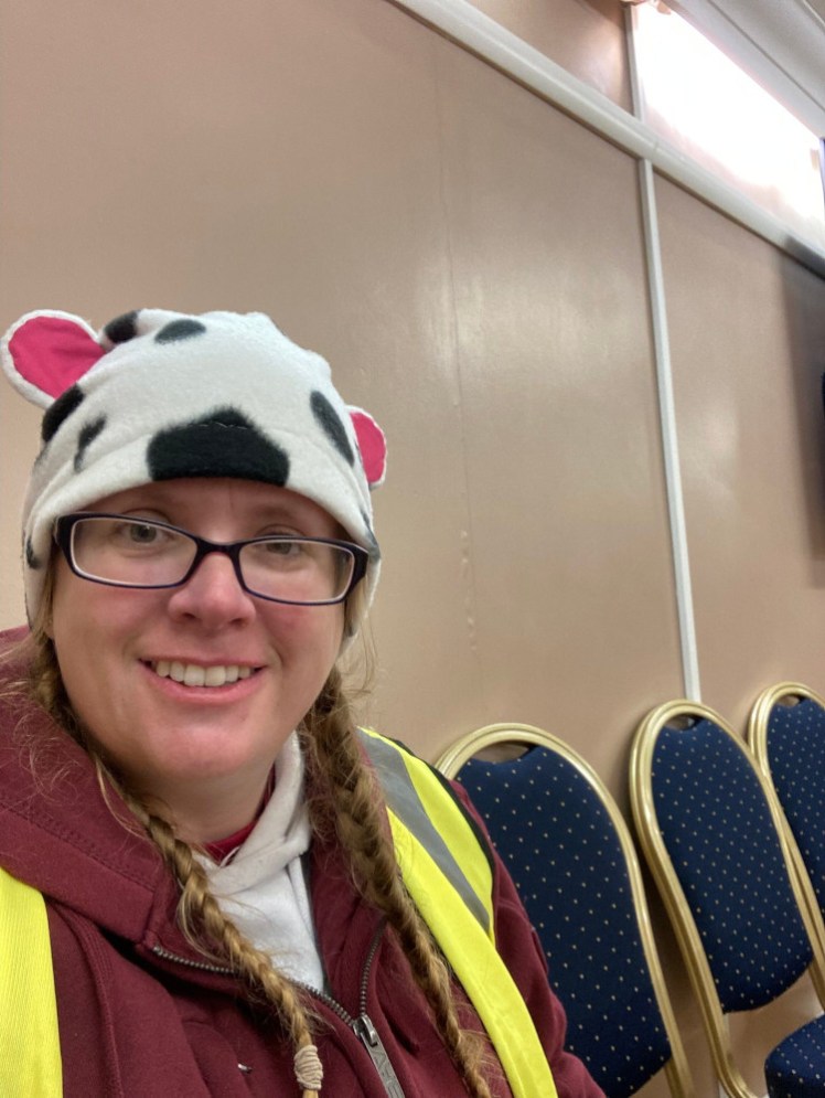 A selfie in the chillout zone against a beige wall. I'm wearing a fleece hat in a cow-pattern, complete with cow ears, a dark red zip hoodie over a white hoodie and a fluorescent jacket over the lot.