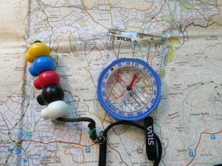 A Silva compass with a blue bezel and a transparent plastic base plate, lying on a map of Dartmoor. Attached to its string is a set of pacing beads, five coloured toggles on a length of cord.