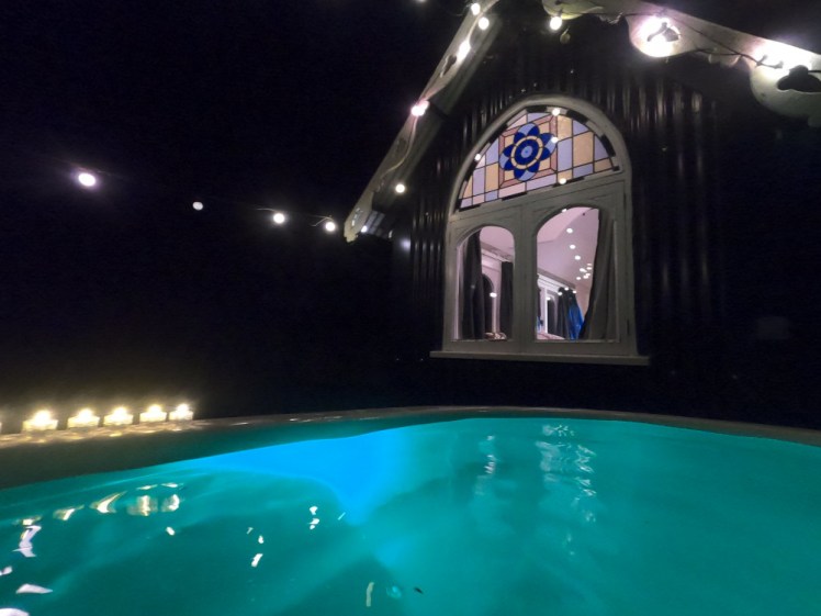 A line of submersible LED tealights on the edge of the hot tub which is lit in light blue. Behind, the stained glass window of the tabernacle glows with the lights on inside and there's a line of lights strung between the hut and a tree.