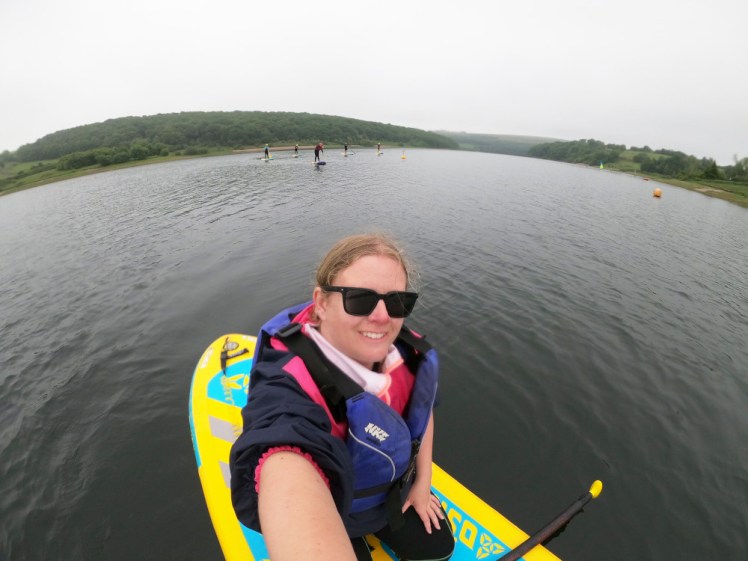 Selfie on a yellow paddleboard. My hair is damp and tied back, I'm wearing a wetsuit and a sailing jacket with pink sleeves under a purple buoyancy aid.