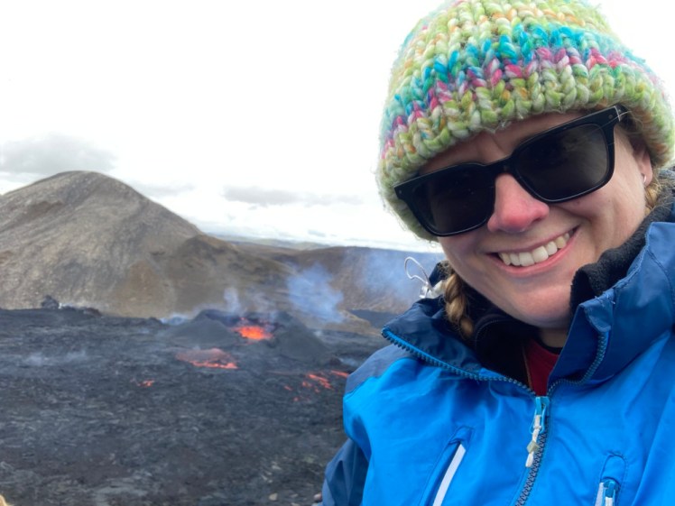 A selfie with a small but erupting volcano in the background. I'm wearing a blue raincoat, a multicoloured bobble hat and a very happy smile.