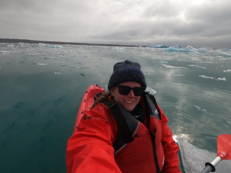 Me in red kit in a red kayak on the glacier lagoon, which is dark turquoise with chunks of ice floating in it.