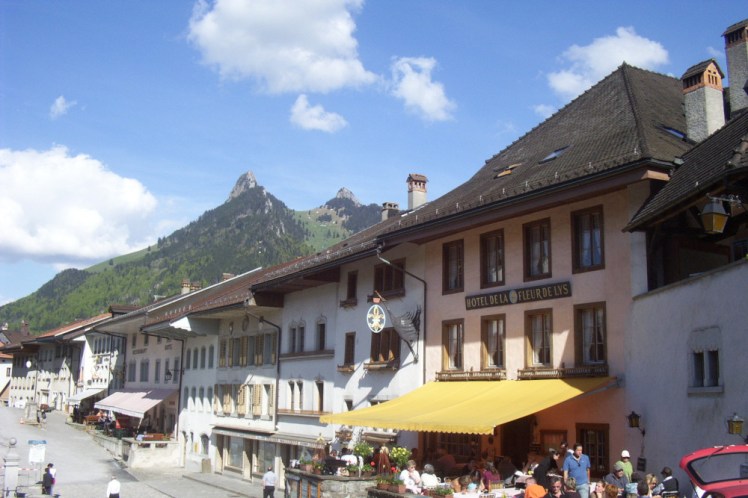 A street in central Gruyeres. Traditional buildings along one side of a cobbled street. The closest, a hotel/restaurant has a yellow awning to keep the bright sun off its patrons. The others are painted white and have heavy wooden window frames and shutters.