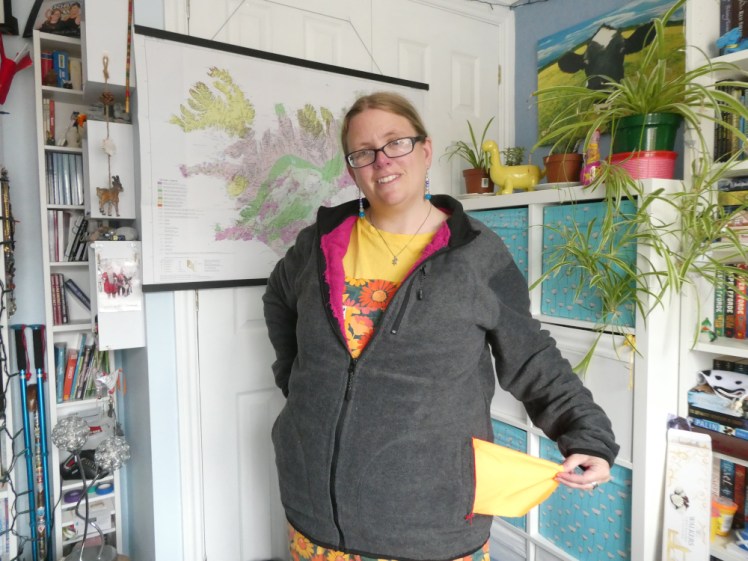 Me in a dark grey fleece jacket with a visible bright pink towel lining visible. I'm pulling out the lining of one of the pockets. It's eye-burning orange but in this light it just looks yellow, like the t-shirt I'm wearing underneath the jacket.