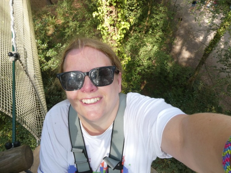 A selfie taken during a high ropes adventure. It's from a high angle so you can see straight down past my face to the ground thirty-odd feet below. I'm wearing a full-body climbing harness, sunglasses and a white Wellies & Wristbands t-shirt