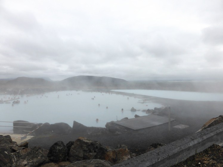 Myvatn Nature Baths, an expanse of blue lagoons under a mist or steam, set among the mountains and with Lake Myvatn just visible right in the background.