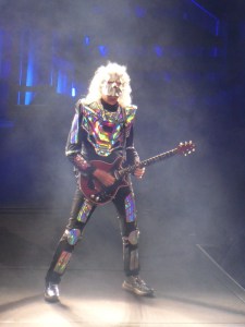 Brian May - a tall man in his mid-70s with huge white curly hair - is holding a red guitar and wearing a black outfit decorated with holographic silver accents. An entire body piece plus plates on arms and legs, matching silver shoes and a silver mask covering his entire face.