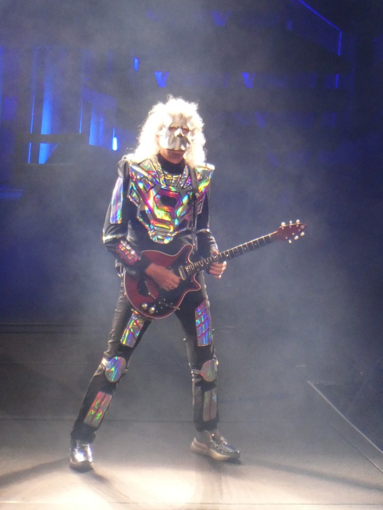 Brian May - a tall man in his mid-70s with huge white curly hair - is holding a red guitar and wearing a black outfit decorated with holographic silver accents. An entire body piece plus plates on arms and legs, matching silver shoes and a silver mask covering his entire face.