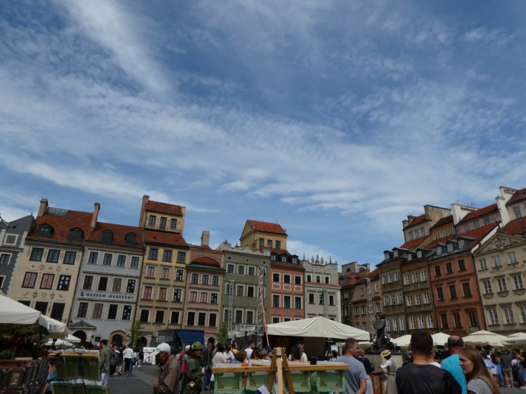 The Old Square in Warsaw. It has the usual tall colourful houses but the middle is buried under umbrellas outside cafes and hundreds of tourists and visitors.