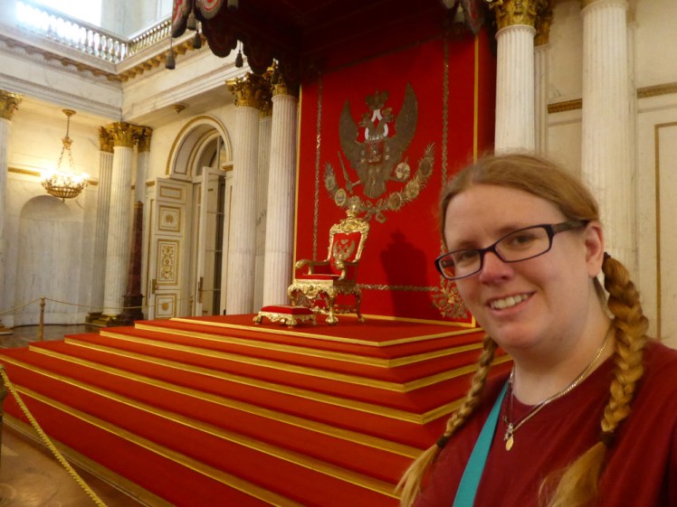 Selfie in a throne room in the Hermitage. The carpet and wall is red and gold, as is the throne, which is really just a dining chair with too much ornate gold on it. I kind of match - my hair is dark blonde with a hint of strawberry in this light, and I'm wearing a red t-shirt.