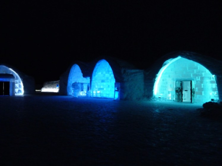 The exterior of the Ice Hotel by night. There are arches lit up in blues and greens.