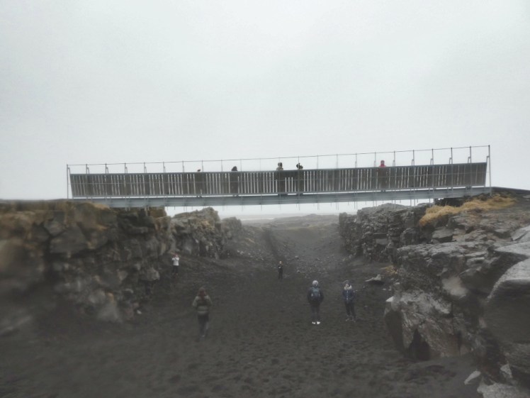 A simple metal bridge stretching between two low blocky basalt cliffs, maybe 20 feet up. There are people standing on the bridge and also in the black dust underneath it. The path underneath slopes up beyond the bridge, taking it back to cliff height within about 100 yards.