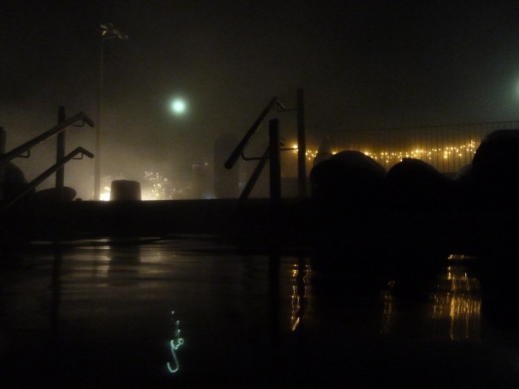 Fontana by night in December 2013. A round green light, possibly the moon, hovers over the scene, which is mostly railings silhouetted against the lights coming up from below, a set of fairy lights illuminating the railings around the raised hot tub and the lights reflecting faintly in the hot water.