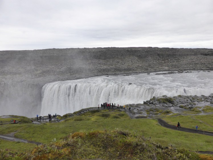 Dettifoss from the west. In the foreground is a hilly grassy area criss-crossed with paths. In the background is a bleak grey rocky desert. Separating the two is a vast curtain of a waterfall.