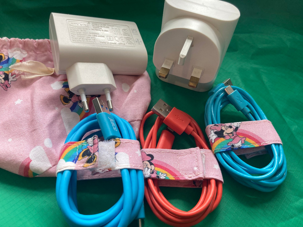 My charging kit - two of the triple USB plugs sitting on top of the flattened pink drawstring bag. Lying in front of them all are two blue wires and a pink one, each coiled and secured by a strip of fabric matching the bag.