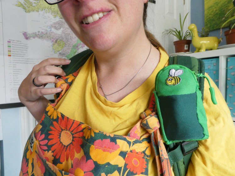 Me from mouth to armpit in the same yellow and orange outfit. I'm wearing the green backpack and on the shoulder strap is a round-topped rectangular pocket, with a dark green lycra front and a bee sewn on the top. It's a bit wonky and a bit rough around the edges.