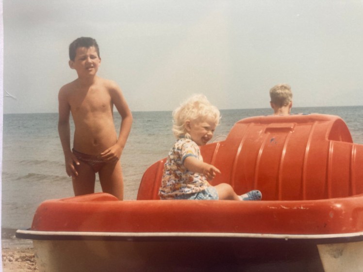 Me in my highly patterned long t-shirt/dress and shorts, sitting on the back of a red pedalo on the beach. Next to me is a boy of about six in a pair of trunks.