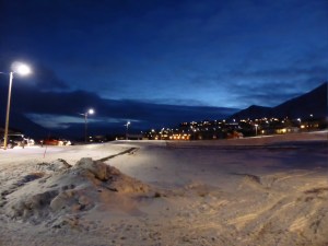 Sky and mountains and the town and snow at about 11am. The sky is dark blue, a little lighter than navy with a hint of light coming in over the mountain. The city is visible under the mountains only because of the lights in its windows. It's still pretty dark.