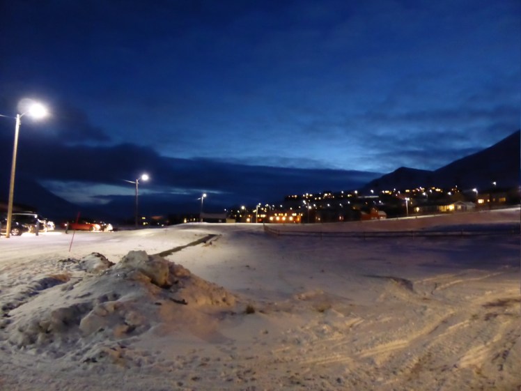 Sky and mountains and the town and snow at about 11am. The sky is dark blue, a little lighter than navy with a hint of light coming in over the mountain. The city is visible under the mountains only because of the lights in its windows. It's still pretty dark.
