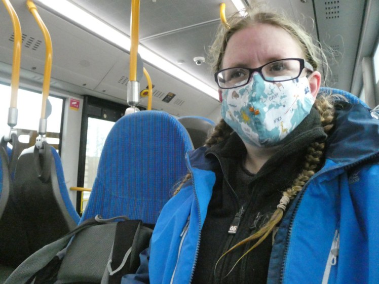 Me on the public bus wearing a mask. I'm wearing a black jacket and blue raincoat. My mask is light blue with a world map, more or less pattern-matched and sprinkled delicately with a few animals.
