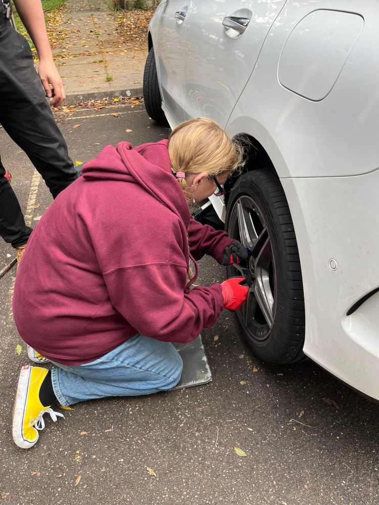 Me. in a dark red hoodie, blue jeans and bright yellow plimsolls, kneeling beside a white car, trying to undo the nuts to remove the wheel.