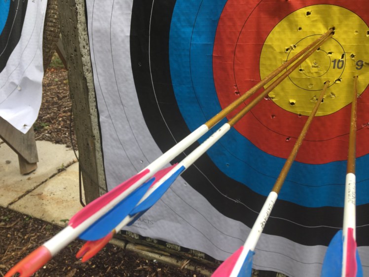 A close-up of an archery target showing four arrows with blue and pink fletchings, all stuck in the gold. This is me practicing while I waited for my pupils to turn up to a taster session and I was very proud of it.