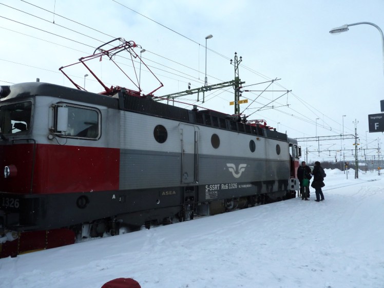 A grey, white and red diesel-electric locomotive in the snow at Kiruna station. It's supposed to go across the border to Narvik but for various reasons, it's going nowhere today.