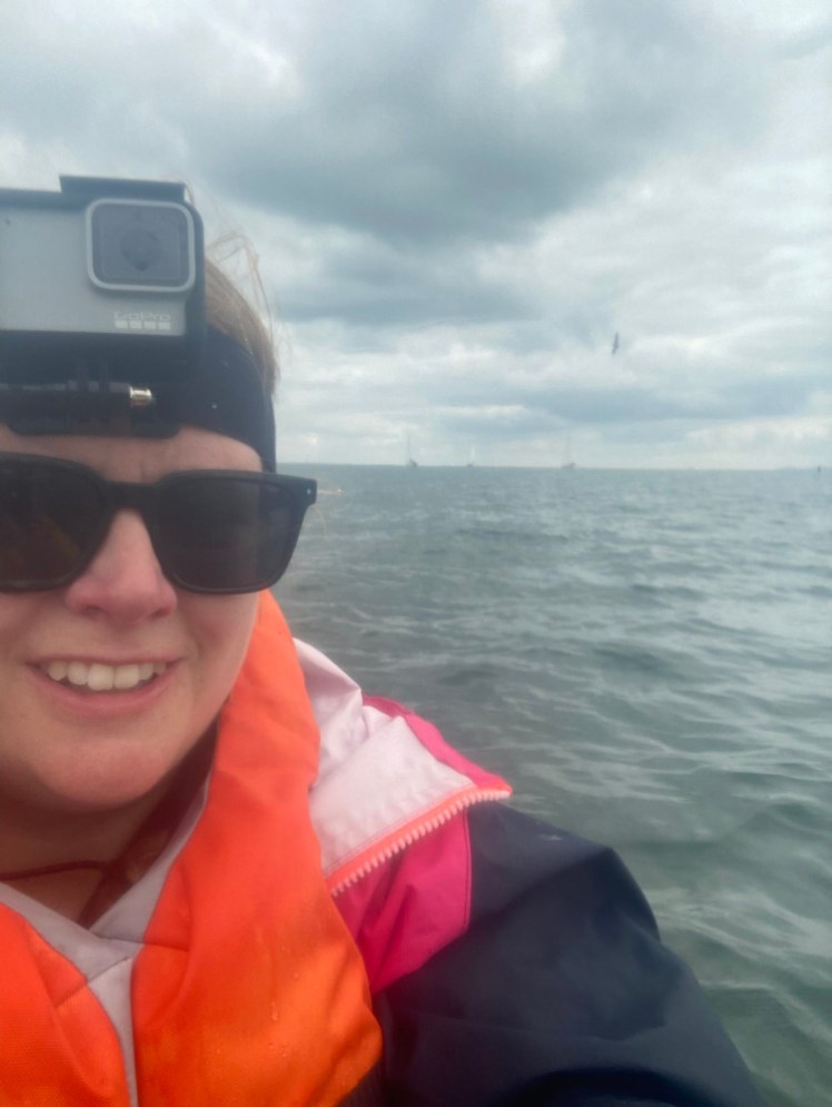Selfie taken on my phone through a misty waterproof case showing me wearing a GoPro on my head, along with an orange buoyancy aid and a navy & hot pink sailing jacket. The day is cloudy and the sea is a kind of blueish grey.