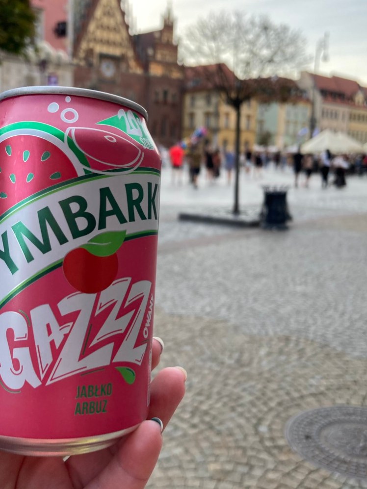 A pink can of something fizzy in apple & watermelon flavour held up against the out of focus background of the colourful square.