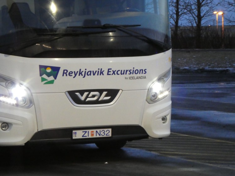 A close-up of the front of a large white Reykjavik Excursions executive coach "bus". You can see the front, the logo, the headlights and about half of the vast windscreen. Behind it, you can just see that it's early evening in winter - the sky is mid-blue and the streelights are bright against it.