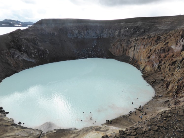 A closer view of the brown muddy crater. The sides are very crumbly and the mud is leaking into the white-blue water. There are lots of people standing on a muddy ledge next to the water and quite a lot of people swimming.