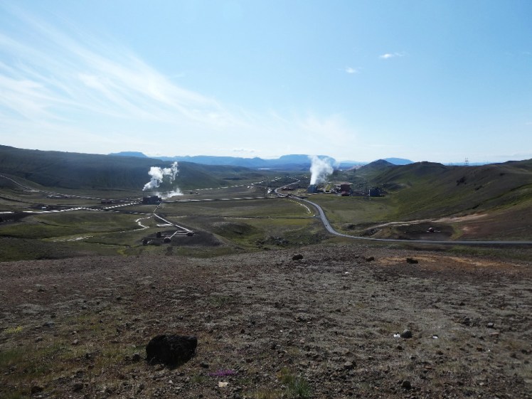 The viewpoint at Krafla, looking into a wide shallow valley criss-crossed with thick silver pipes, with a geothermal power station sending up clouds of steam.
