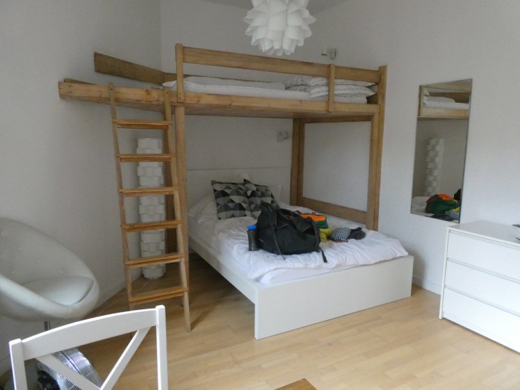 My apartment in Wrocław, a studio where I'm standing in the kitchen with my back to the units and the picture is of the bed, a plain Ikea bed with a hand-made wooden frame creating a bunk bed above it. Everything is very white except the wooden frame and the light laminate flooring.