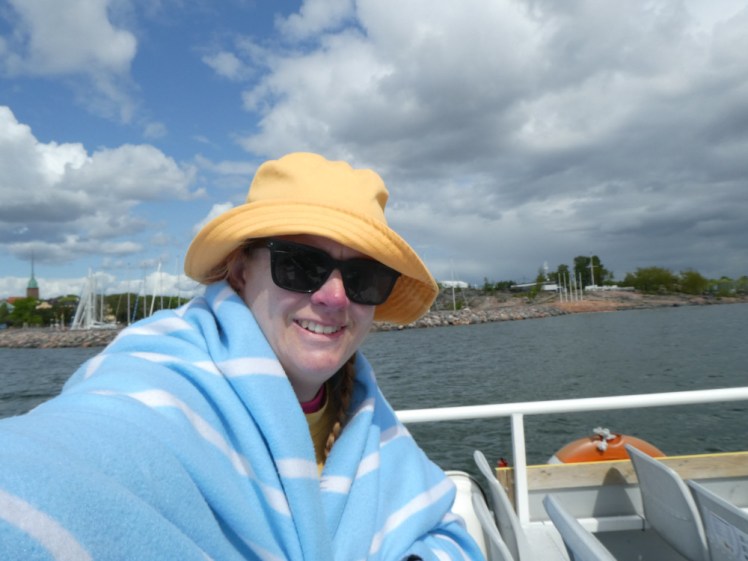 A selfie on a boat, wearing a yellow hat and wrapped in a light blue blanket. Behind me, you can see Helsinki and also a threatening cloud coming over.