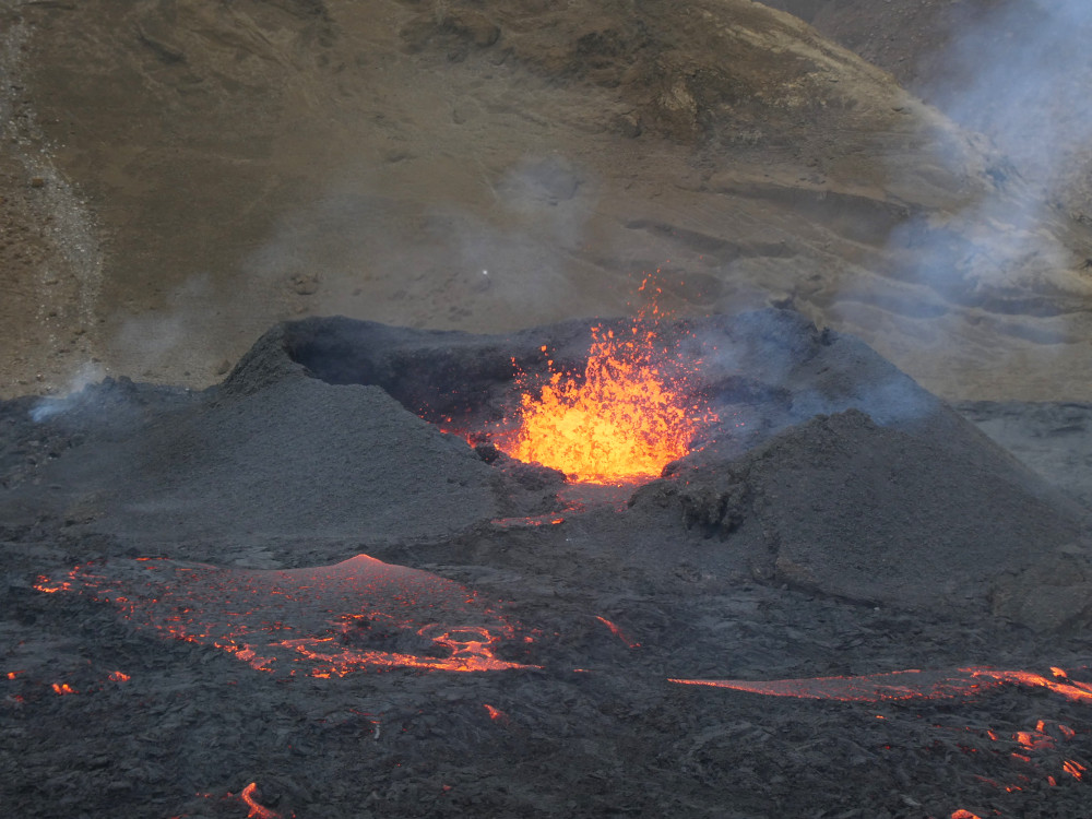 A small black volcano with a yellow-orange explosion of flying fire in the middle. Black lava with orange hot edges is oozing down in front.