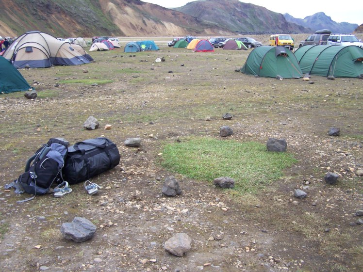 A campsite in Iceland. The background is just small tents of various shapes and colours with streaky mountains right at the back. In the foreground is the patch of grass surrounded by rocks where I camped the night before. My luggage is packed next to it: a 45l backpack, a 100l black duffle bag and a pair of blue sandals which I've opted not to pack, for some reason. Later, I returned to this spot and put my tent back up when I discovered the bus I expected wasn't running that day.